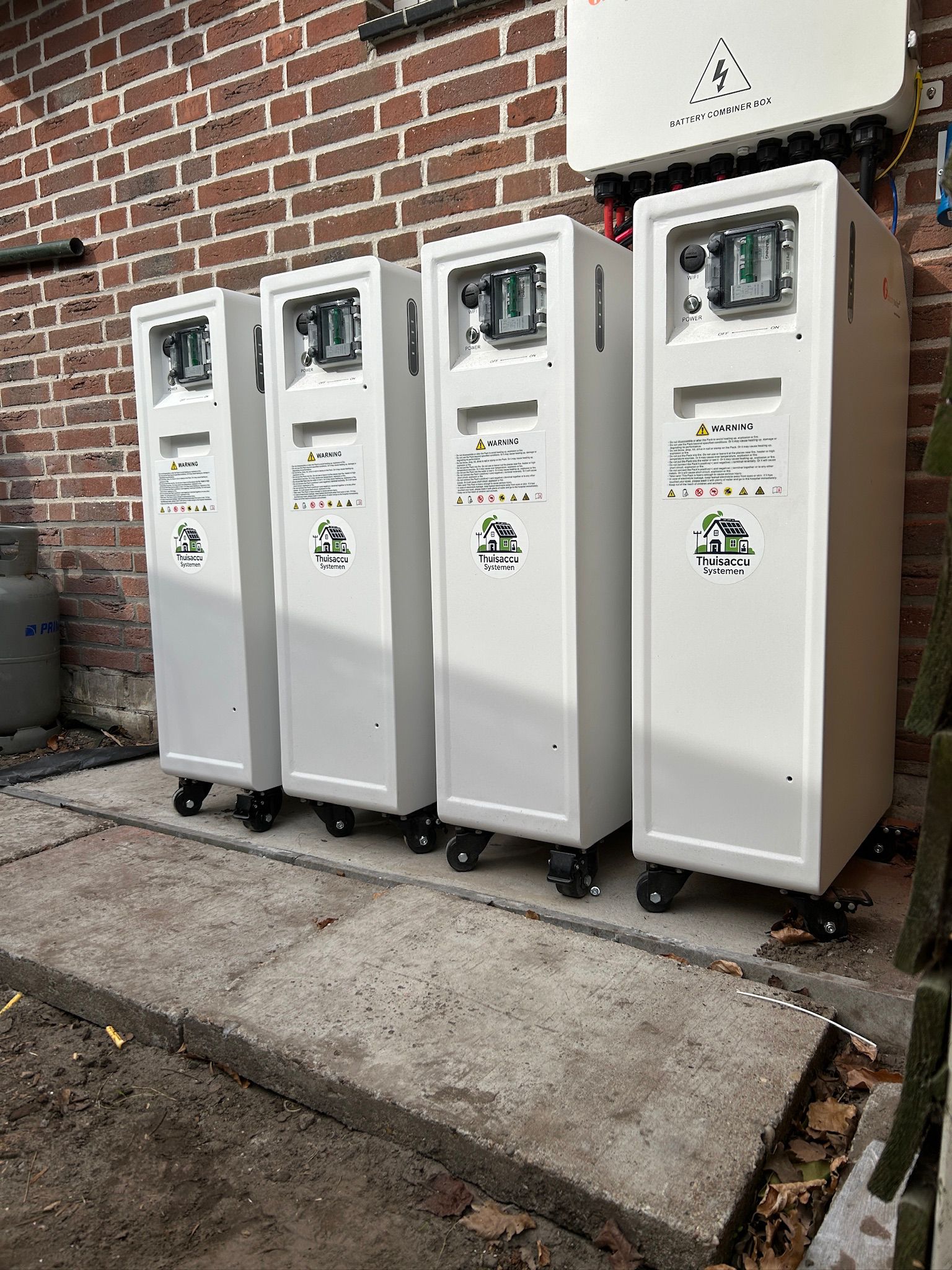 Vier LiFePO4 thuisaccu's naast elkaar tegen een bakstenen buitenmuur